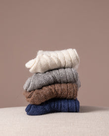 Stack of four pairs of socks in white, grey, brown, and blue on a beige surface with a beige background.