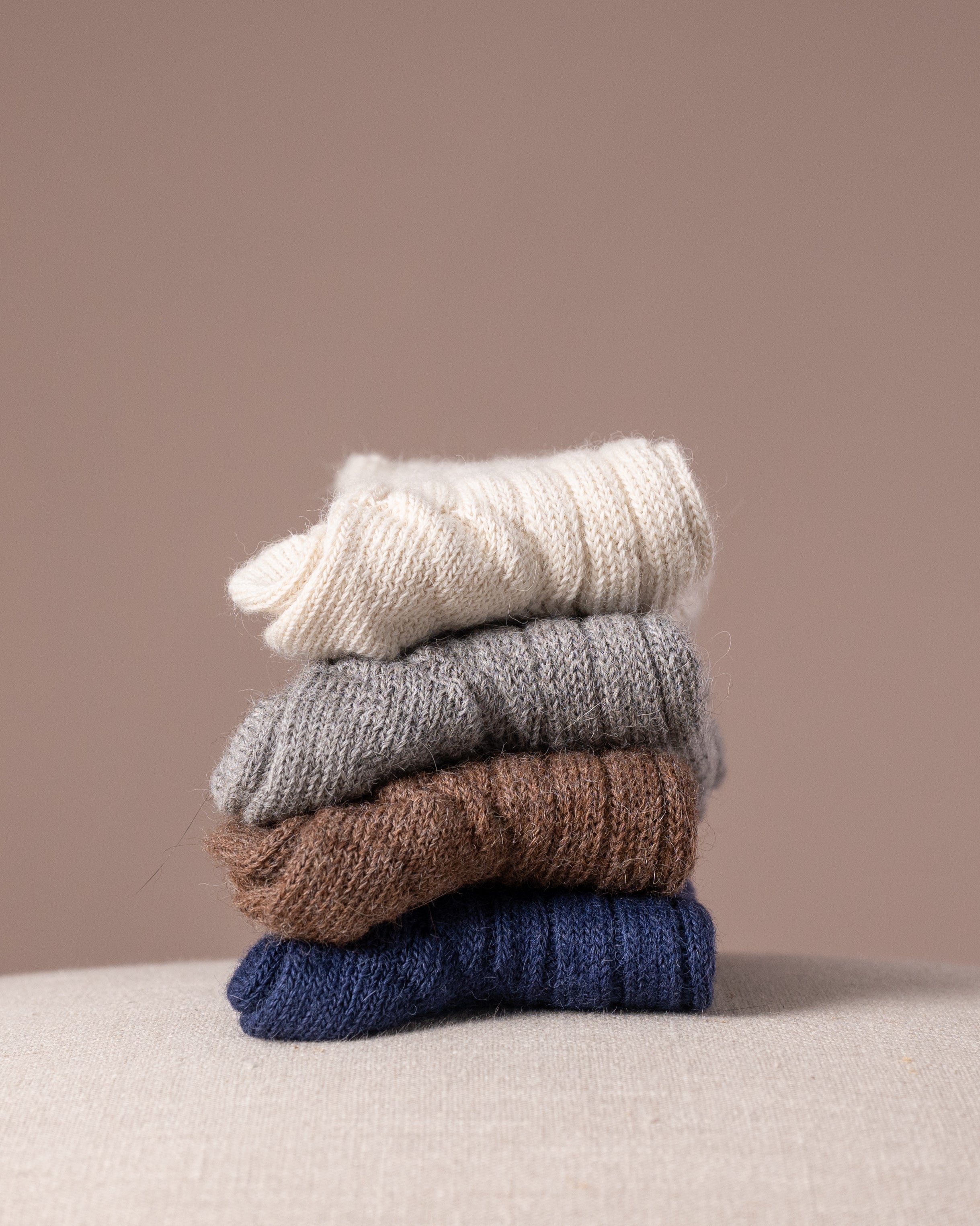 Stack of four pairs of socks in white, grey, brown, and blue on a beige surface with a beige background.