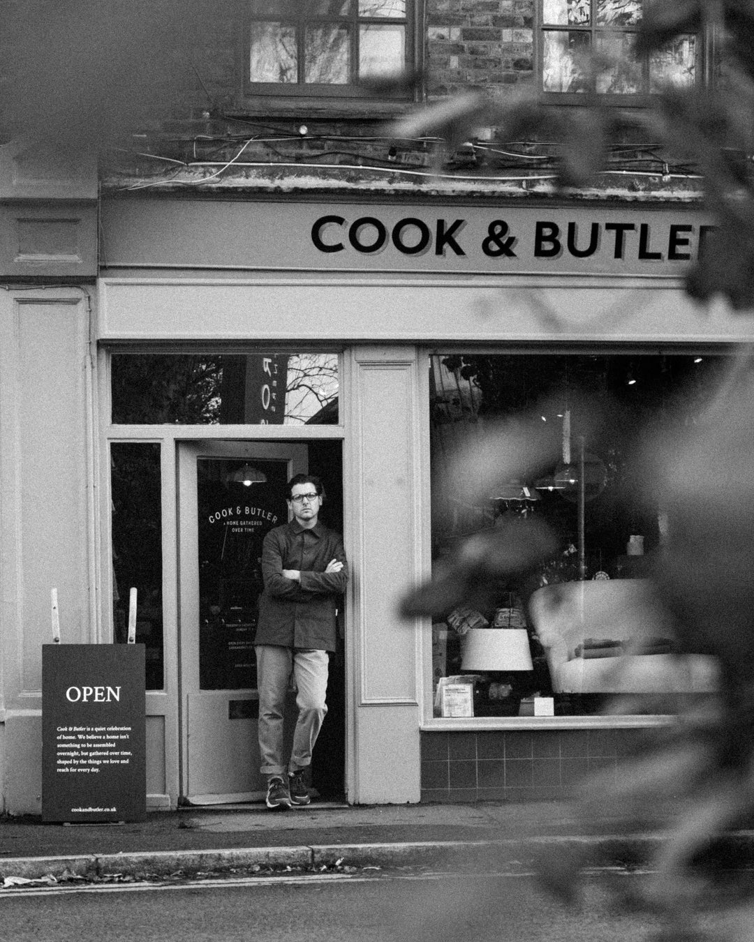 Shop keeper standing outside a shop named 'Cook & Butler' with 'OPEN' sign.