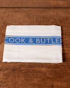White towel with blue 'Cook & Butler' stripe on a wooden surface