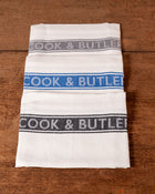 Folded white tea towels with 'Cook & Butler' text on a wooden surface