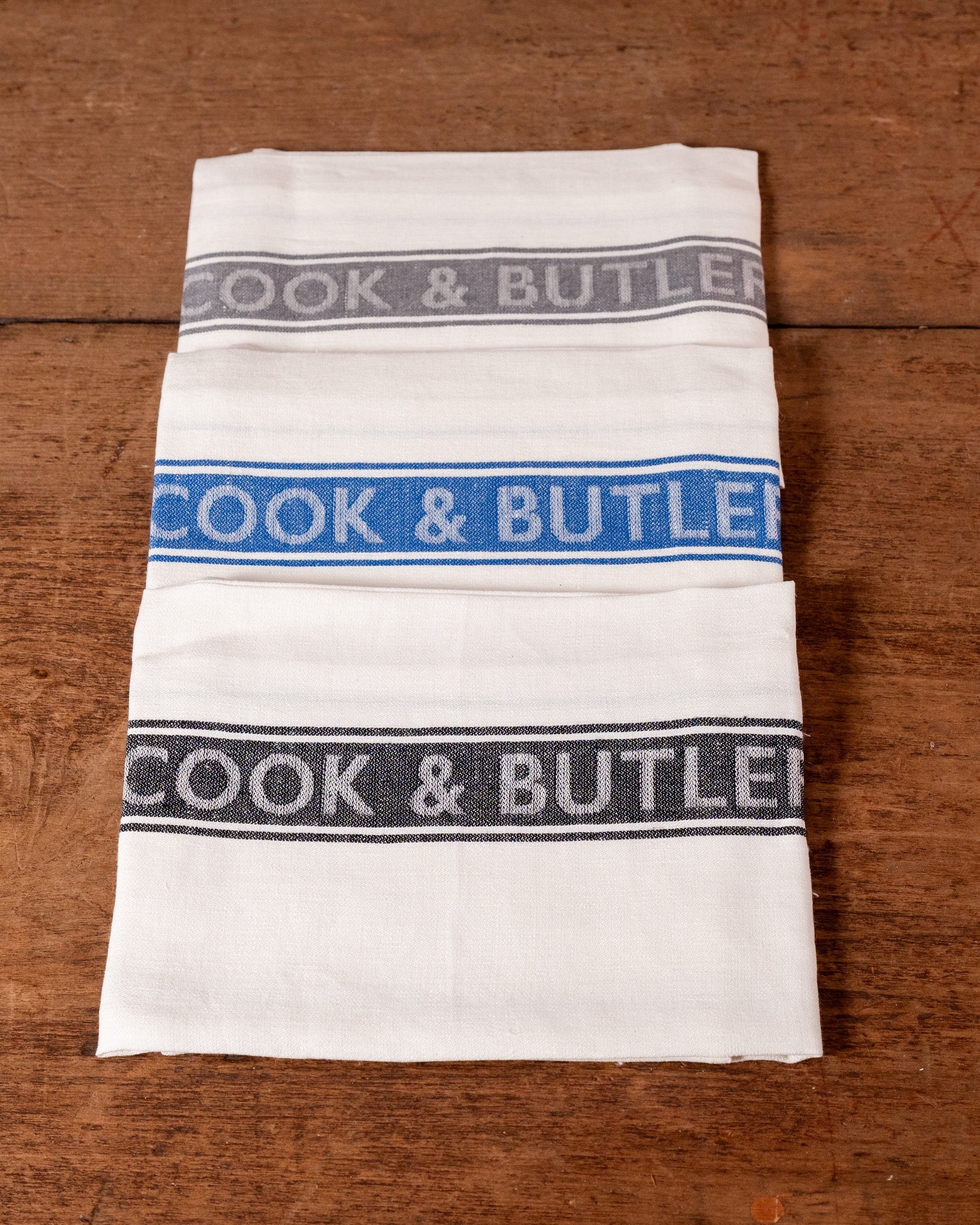 Folded white tea towels with 'Cook & Butler' text on a wooden surface