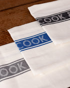 White tea towels with 'COOK & BUTLER' branding in different colors on a wooden surface