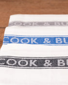Stack of white tea towels with 'Cook & Butler' branding on a wooden surface