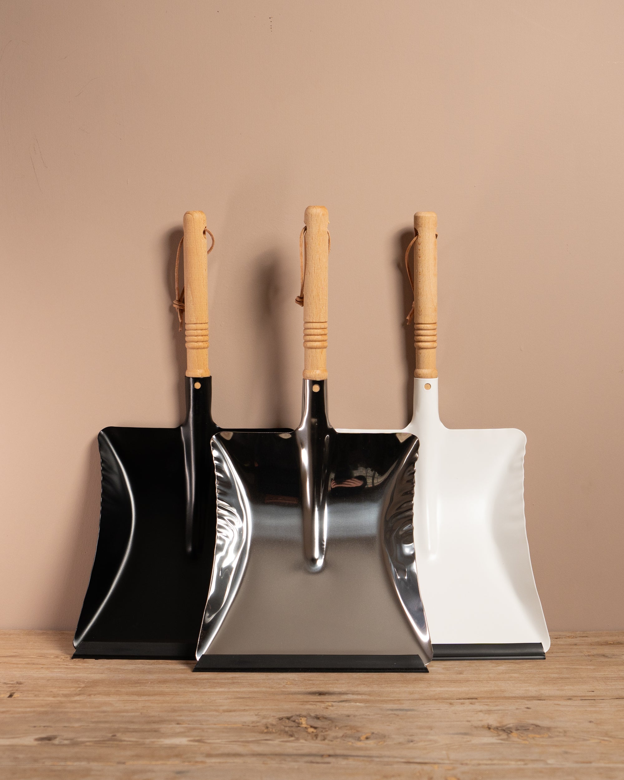 Set of three dustpans with wooden handles on a wooden surface against a beige wall.