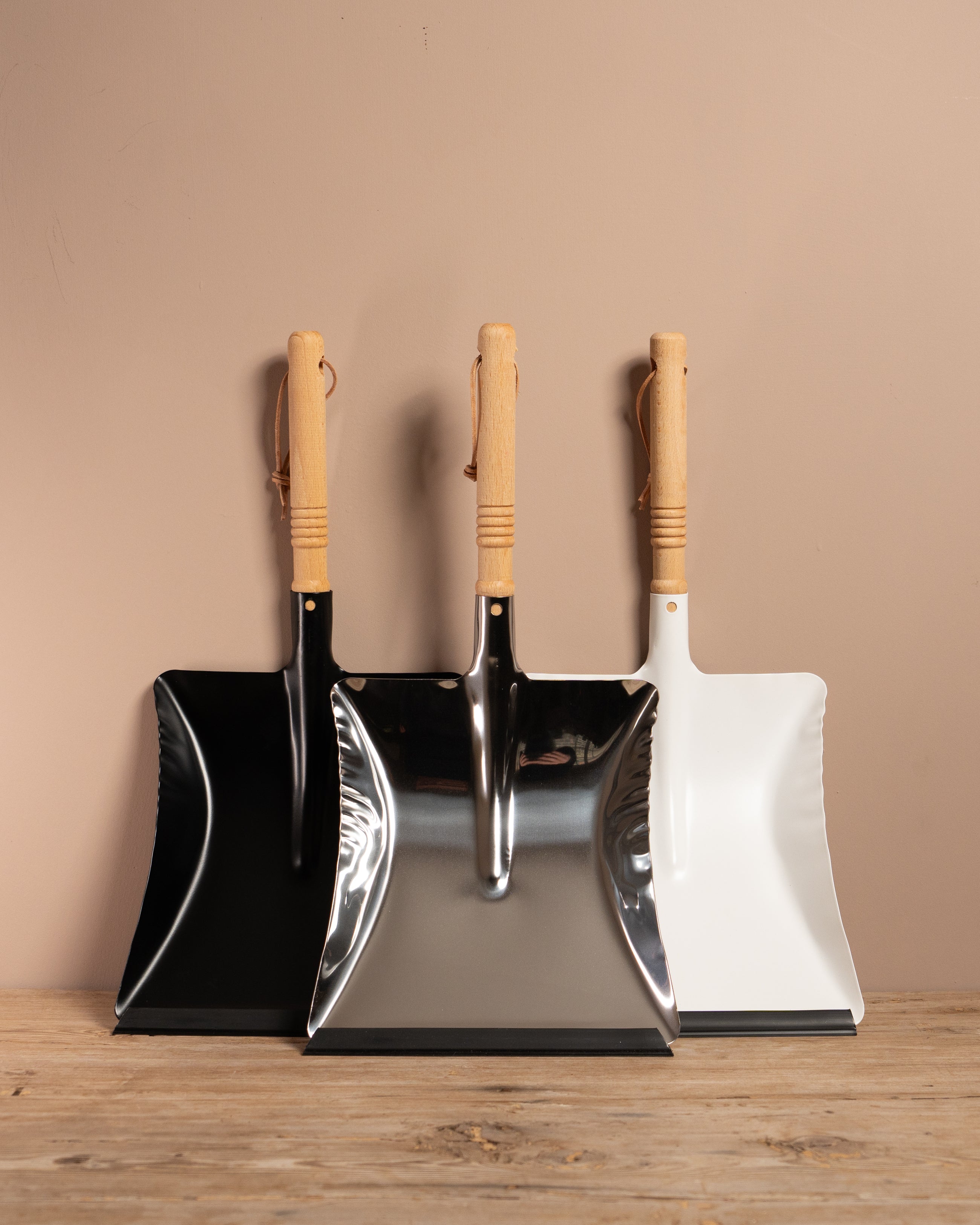 Set of three dustpans with wooden handles on a wooden surface against a beige wall.
