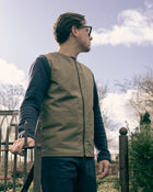 Man wearing a beige vest and glasses standing outdoors with a cloudy sky.