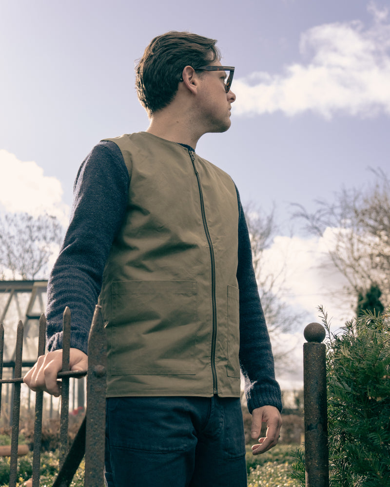 Man wearing a beige vest and glasses standing outdoors with a cloudy sky.