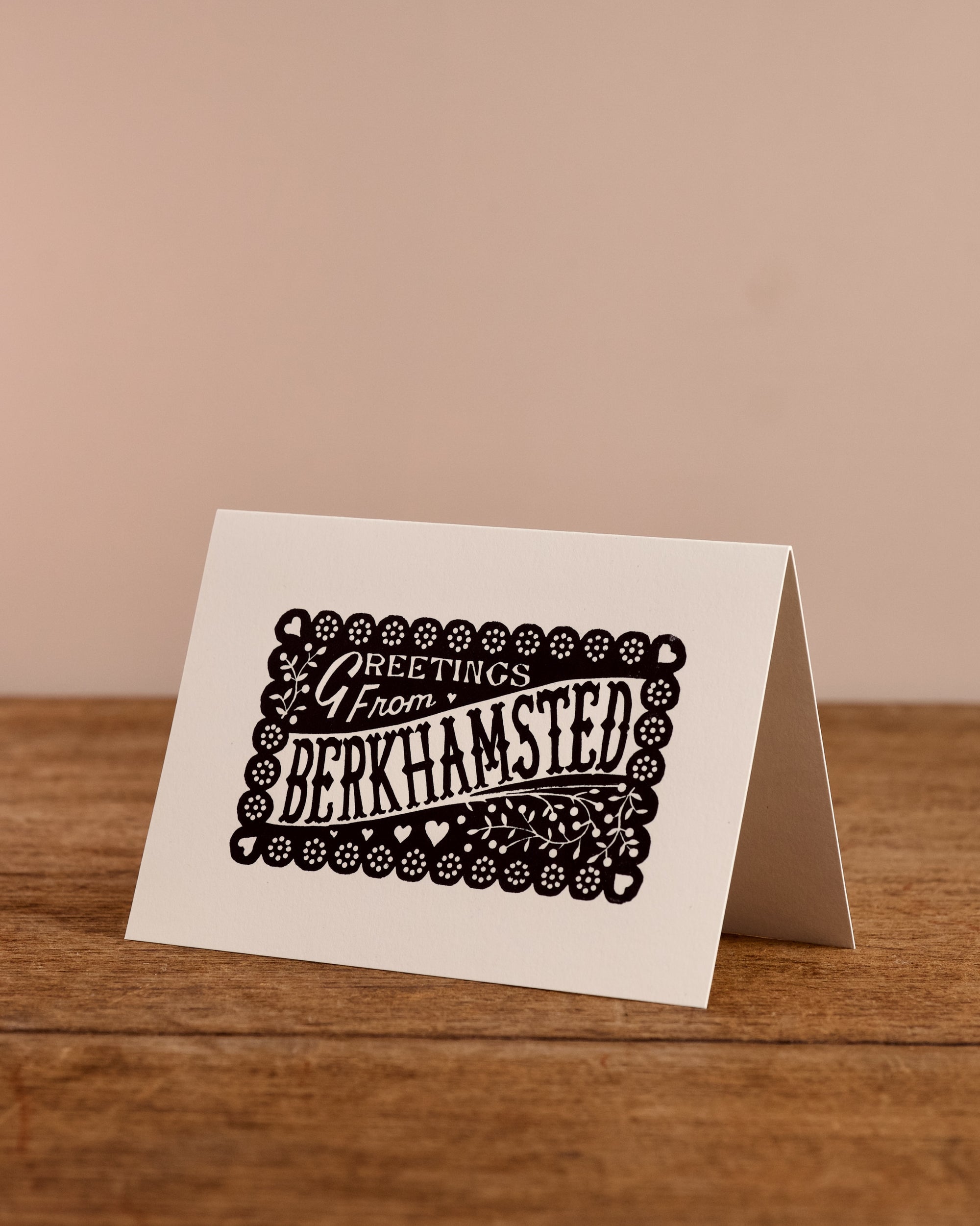 Greeting card with 'Greetings from Berkhamsted' text on a wooden surface and beige background