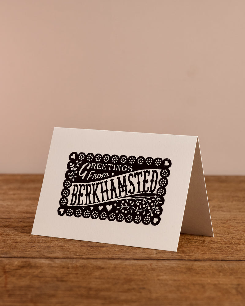 Greeting card with 'Greetings from Berkhamsted' text on a wooden surface and beige background
