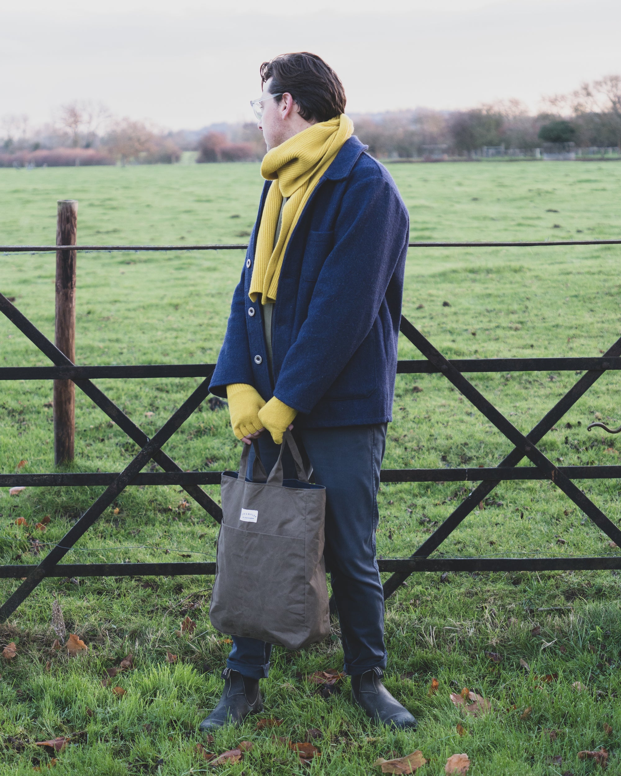 Man standing in a field holding a bag, wearing a blue coat and yellow scarf.
