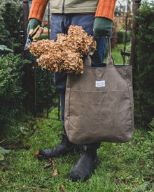 Person holding a brown tote bag filled with dried plants in a garden setting
