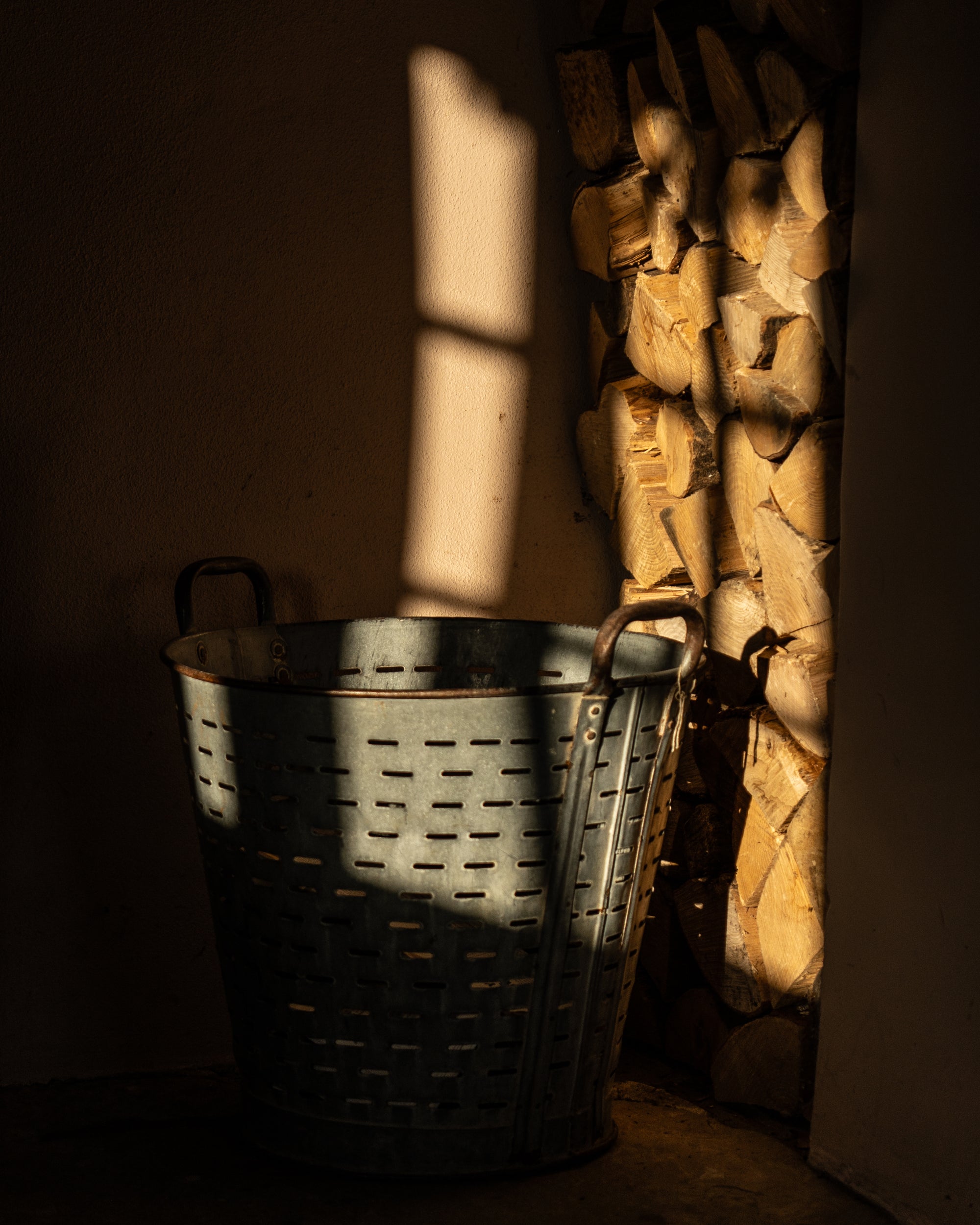 Metal olive basket next to some stacked firewood being lit by sunlight