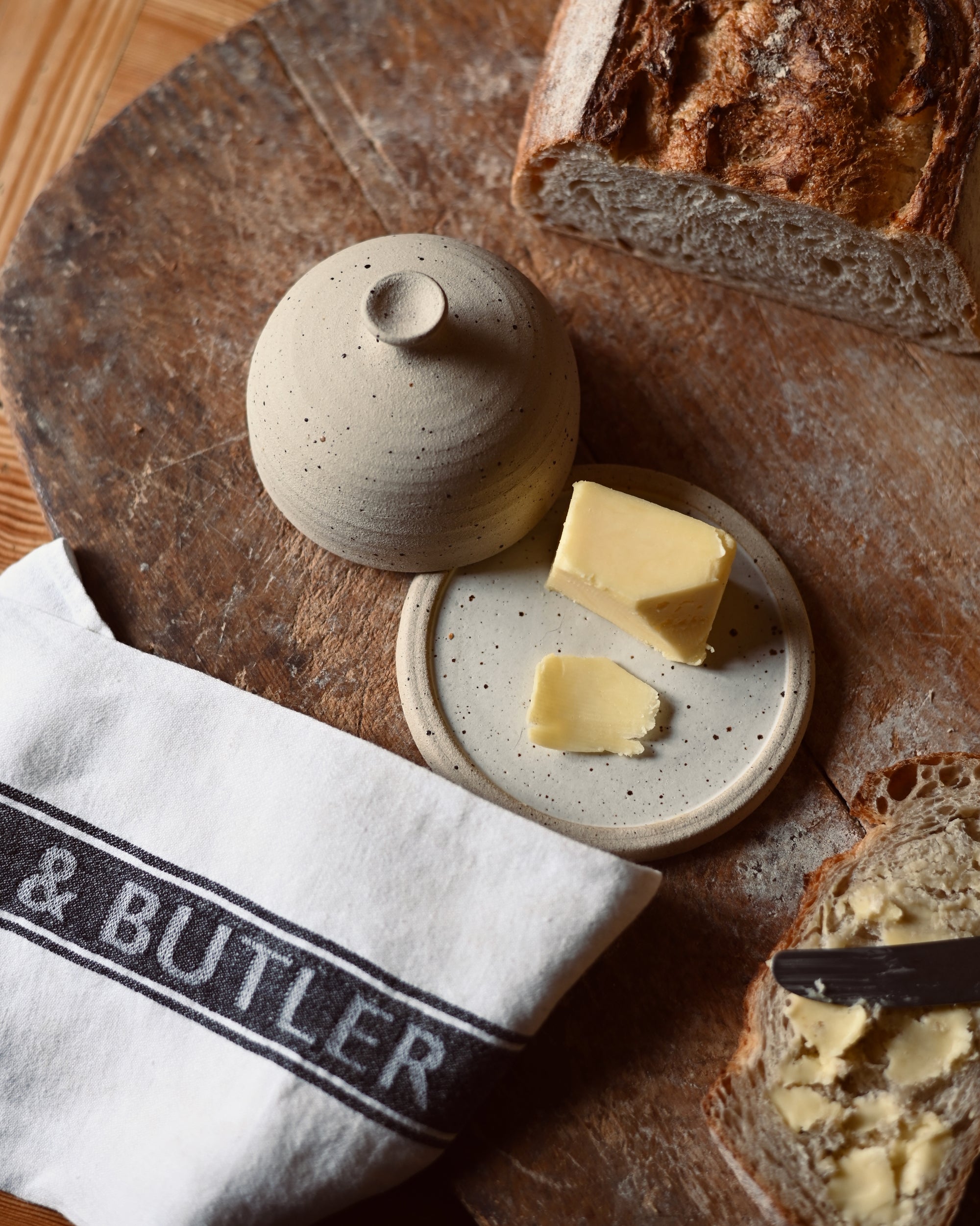 Butter dish with butter on a wooden board with crusty bread, next to a 'Cook & Butler' branded tea towel.