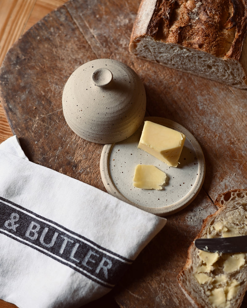Butter dish with butter on a wooden board with crusty bread, next to a 'Cook & Butler' branded tea towel.